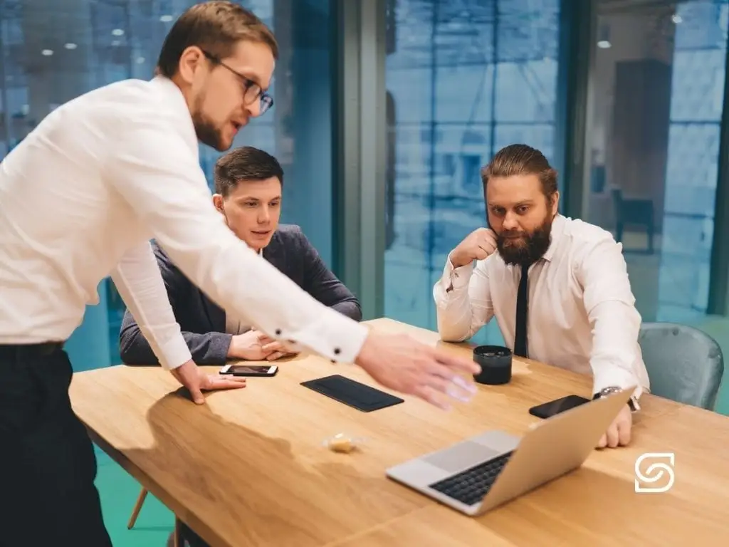 Three men in shirts and ties gathered around a laptop at a wooden meeting-room table, one standing and gesturing while explaining something.