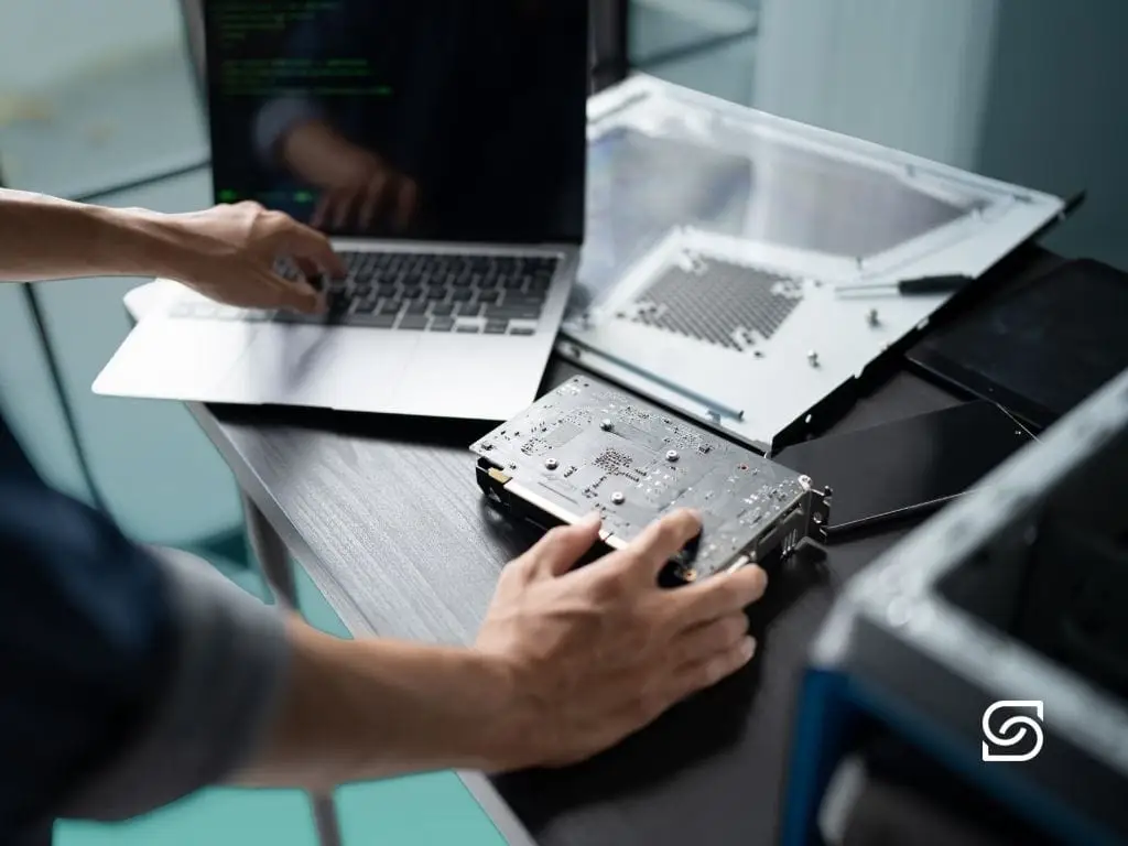 Overhead photo of a technician's hands repairing a laptop with the screen removed, holding a circuit board next to the disassembled chassis.